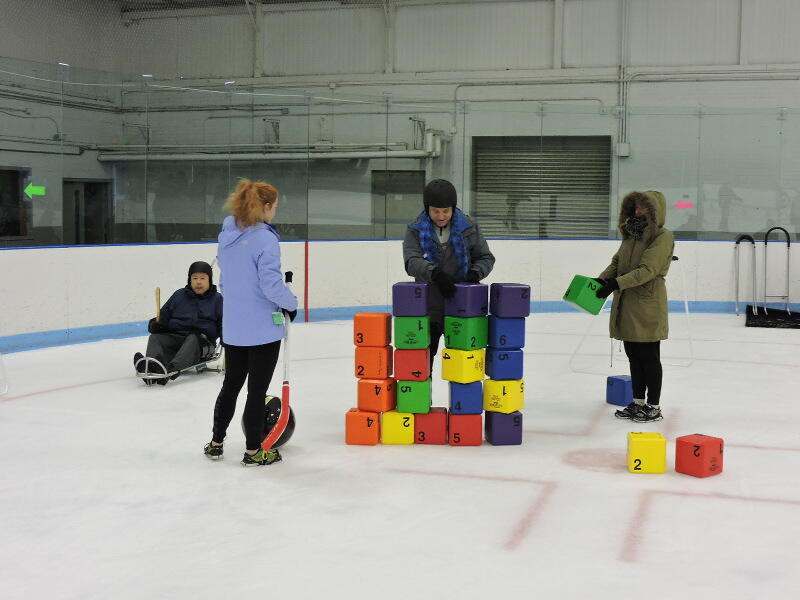 Two people are stacking colorful blocks on the ice. Another person is waiting nearby with a hockey stick and a large ball. Behind them, a man is skating using an ice sled.