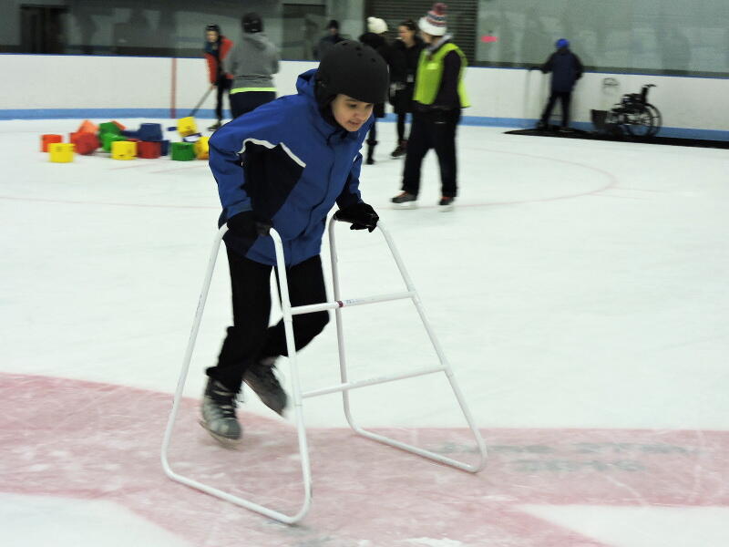 A young person wearing a helmet is skating using a skate walker. Behind them, a group of people is looking at colorful blocks on the ice. One of the people is using a hockey stick to play with a block.