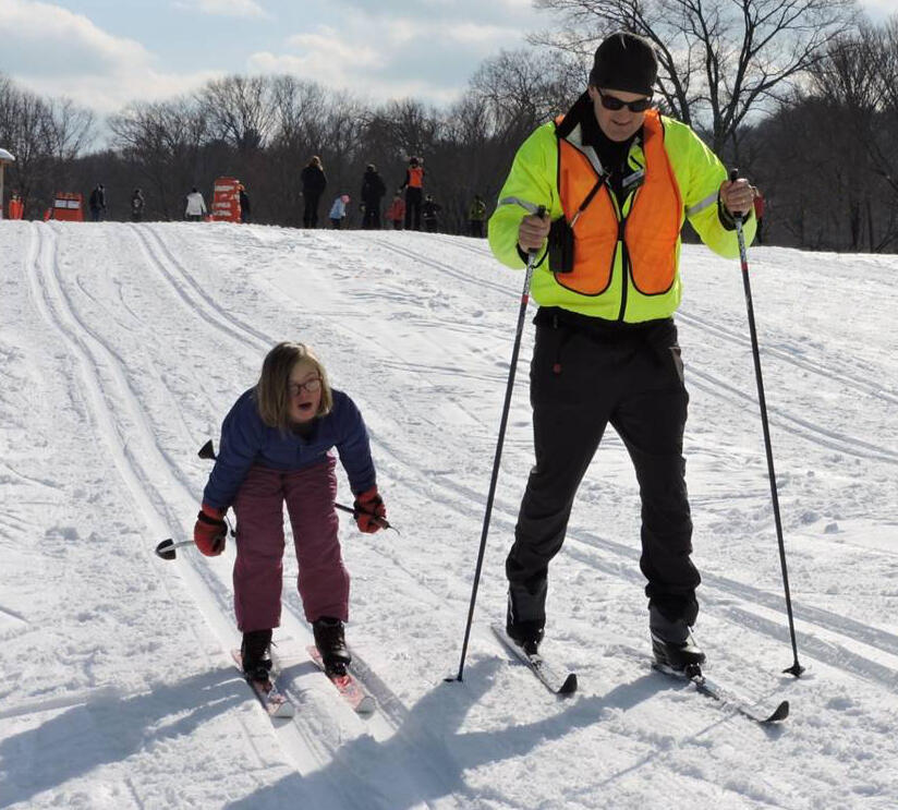 A man and a girl are skiing down a gentle hill next to each other. The girl is leaning forward with her poles stretched out behind her. She is smiling.