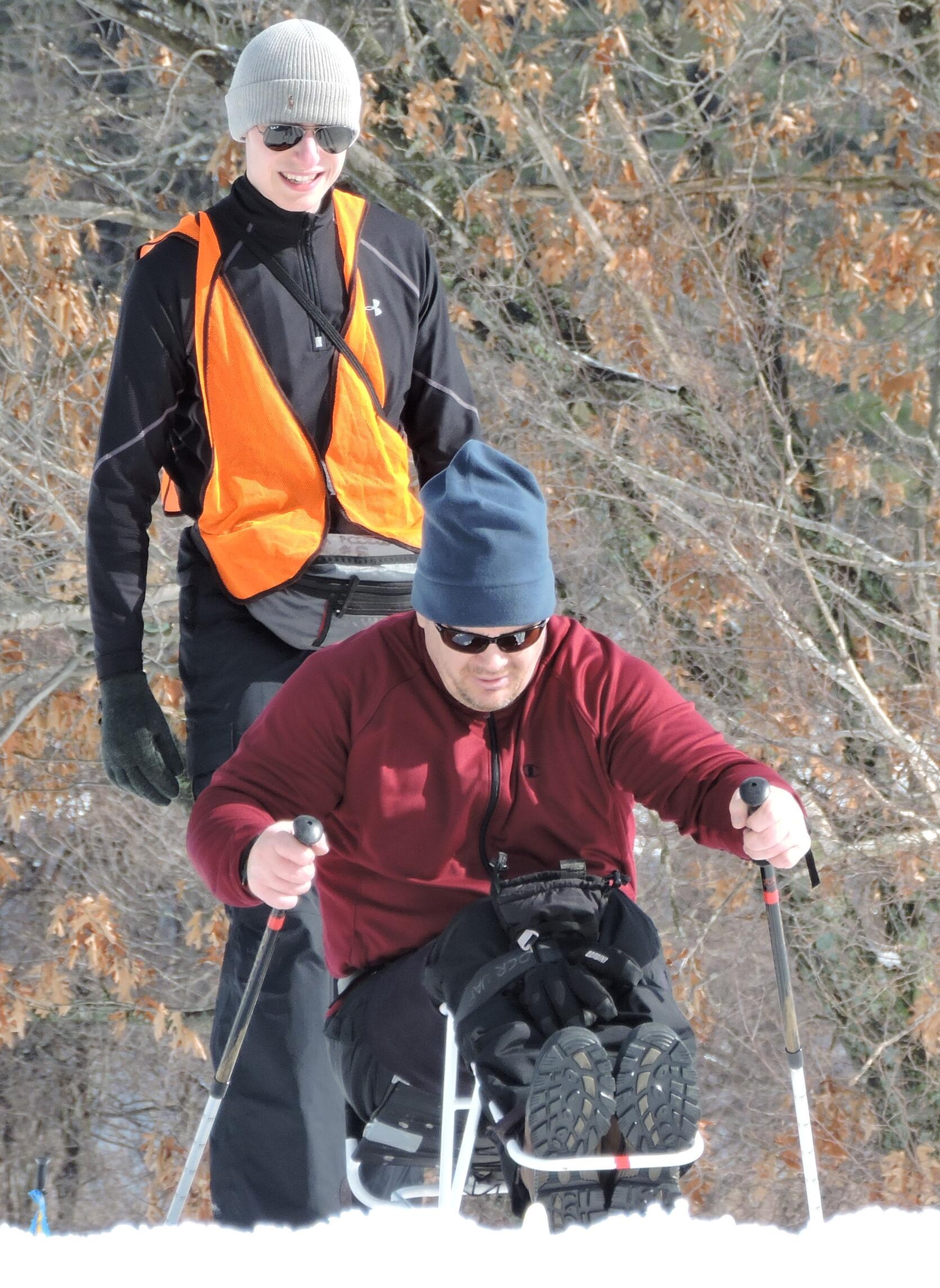 A man in a sit ski is pushing himself up a slight hill using two short ski poles. His knees are bent, and a strap is holding his legs to the sit ski frame. Another person is walking behind him.