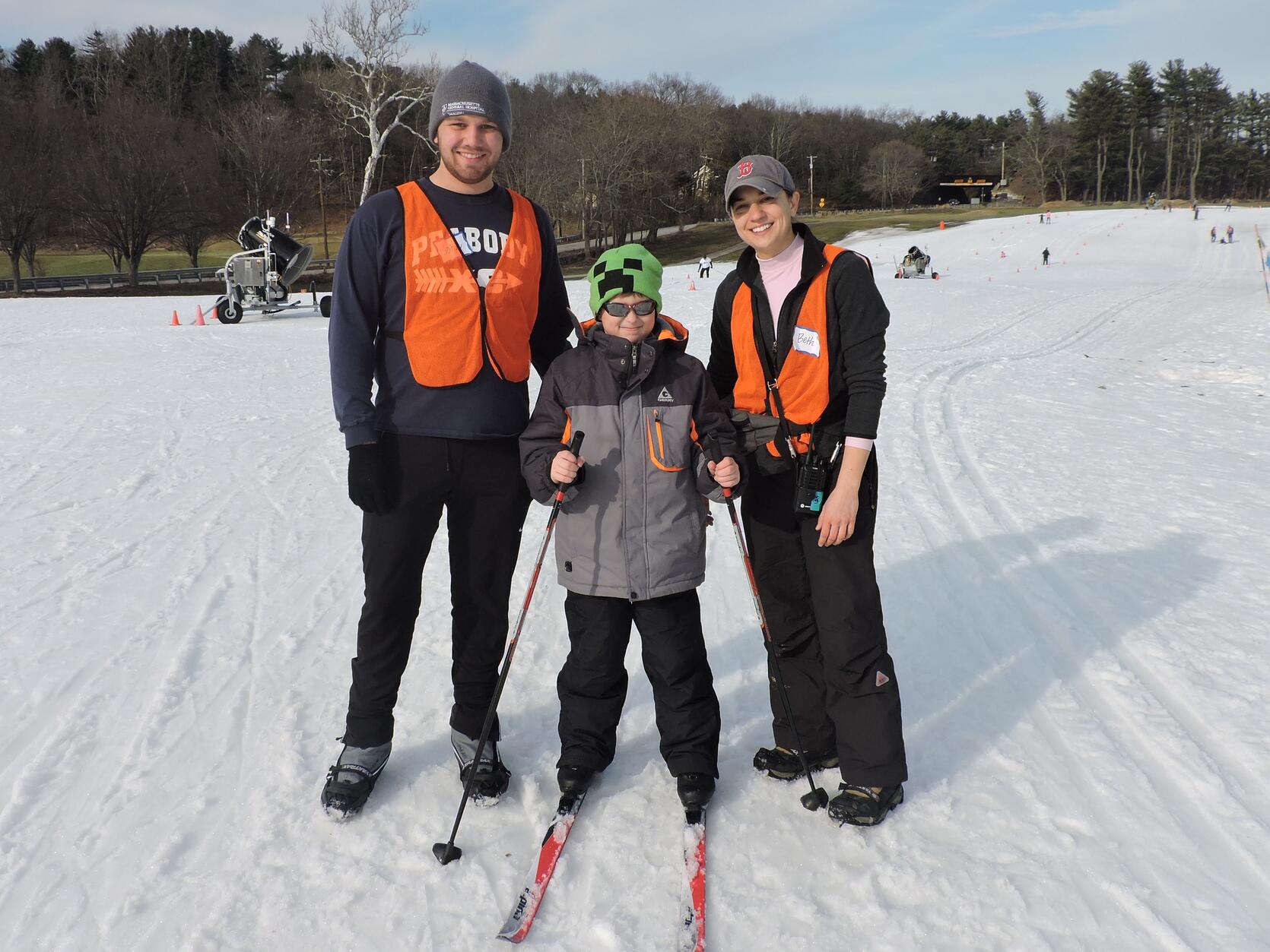 A young boy dressed in warm winter clothes is using cross-country skis and ski poles on the groomed trail. He is standing and smiling at the camera. There are two other people, one on either side of him, wearing ice grippers on their boots. These two individuals are also smiling.