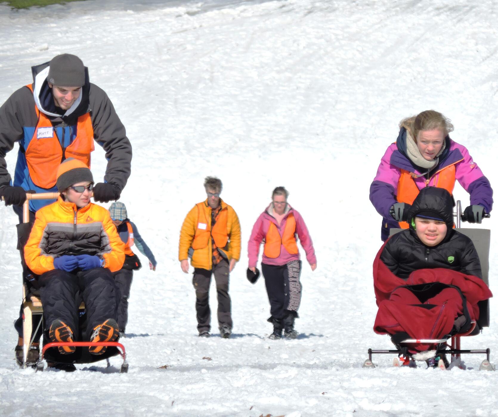 A kicksled user and a sit-skier are being pushed down the hill by volunteers. Behind them, two other people are walking down the hill wearing ice grippers on their shoes.