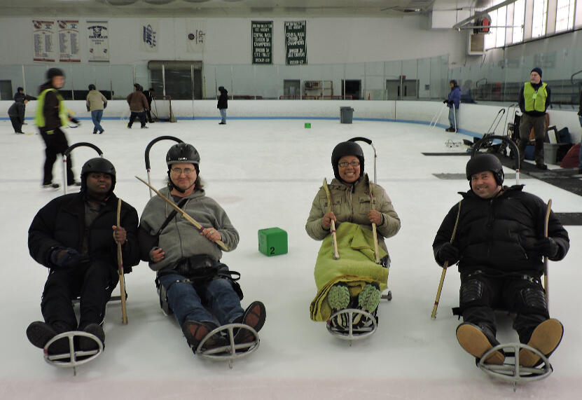 Four skaters in ice sleds are holding short hockey sticks and smiling at the camera. Behind them, other skaters are taking shots at a hockey net.