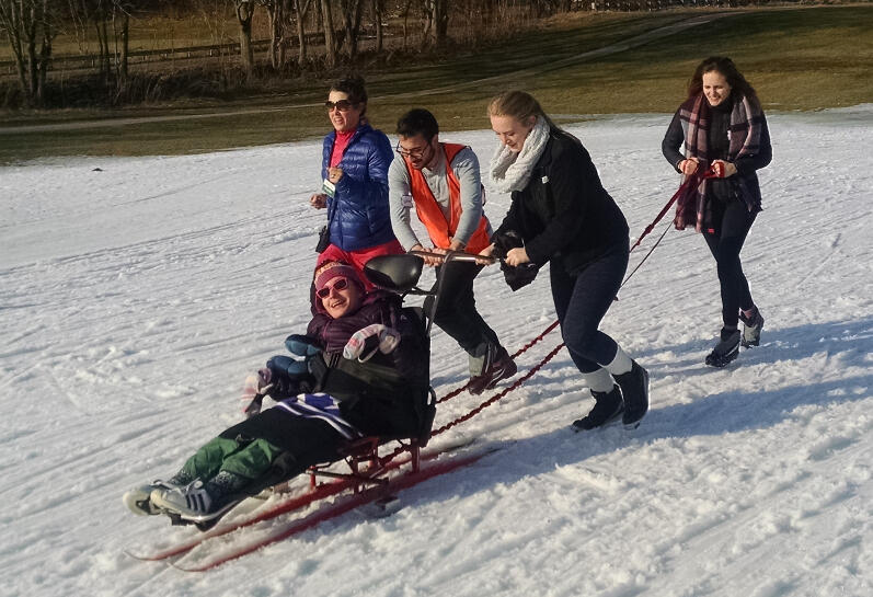 A smiling woman in a sit ski is being pushed up a slight hill by two people behind her. The woman is sitting with her legs straight out in front of her, and is wrapped in a blanket. A third person is holding a tether that has been attached to the sit ski, and a fourth person is jogging alongside.