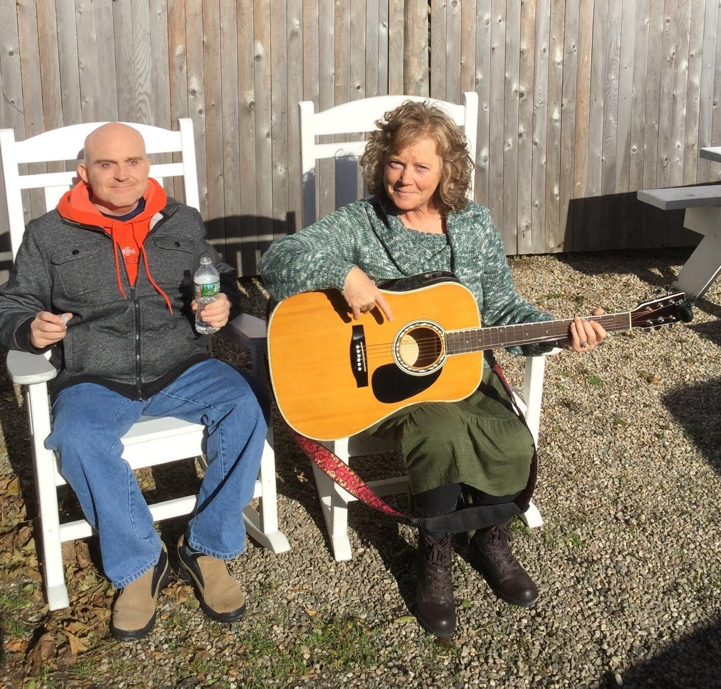 Cheryl and Glenn are in rocking chairs outdoors with Cheryl playing guitar