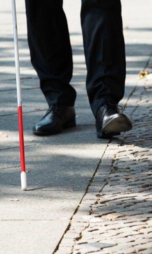 Individual walking on sidewalk in black pants and black shoes using white cane to navigate safely
