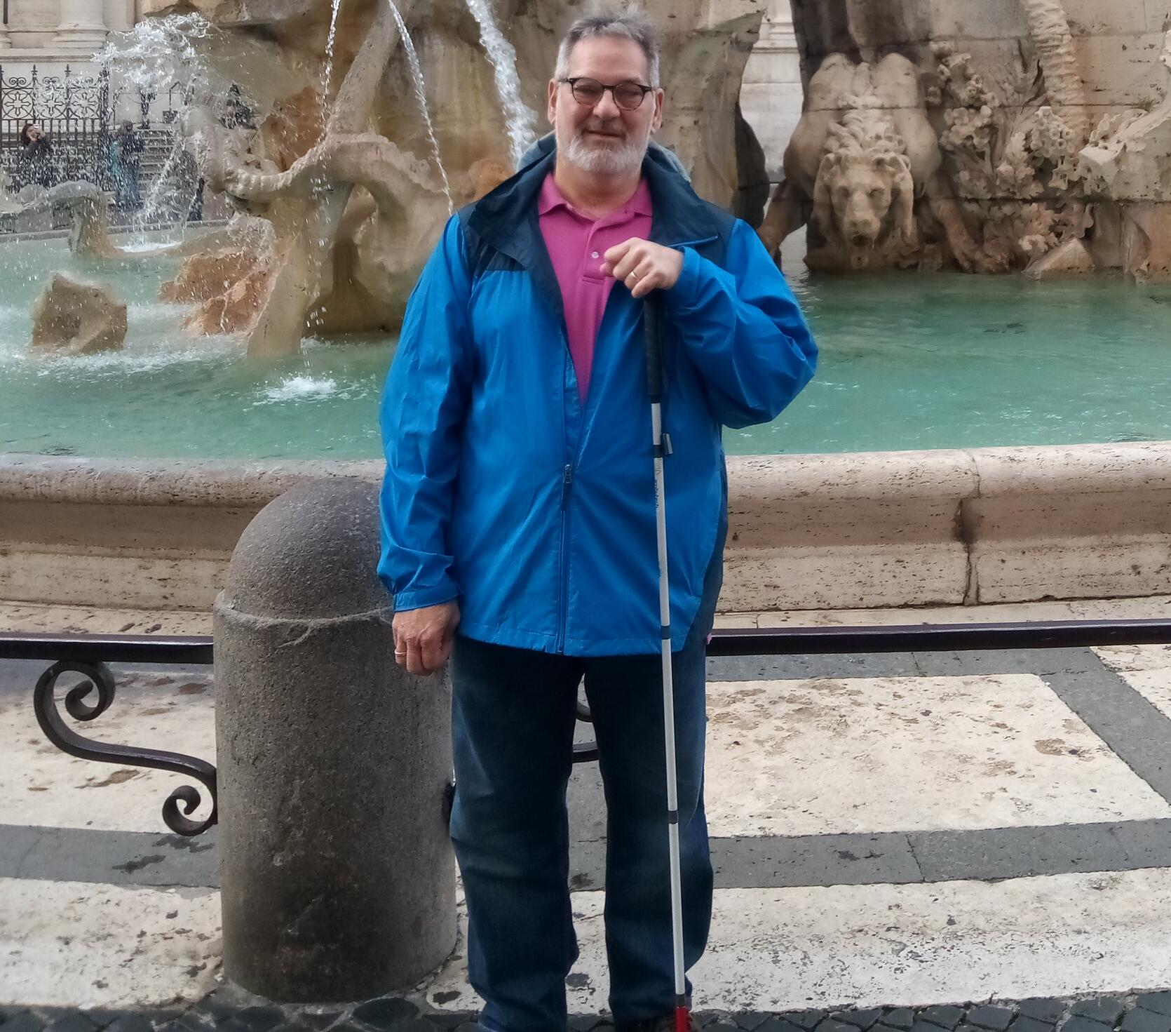 MCB Region 4 Director Alan White standing in blue jacket with white cane in front of fountain