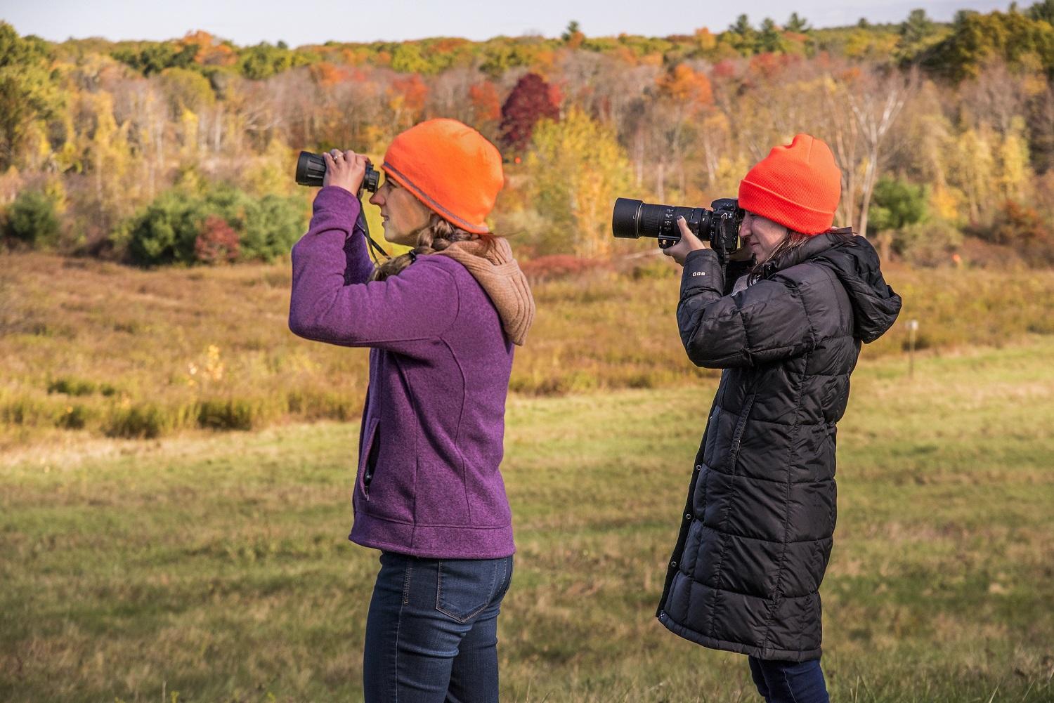 Two women wear orange hats while birdwatching and photographing wildlife in fall. 