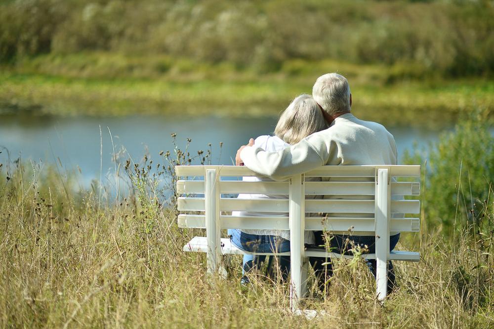 Elderly man and woman embrace while sitting on a white bench together overlooking a pond