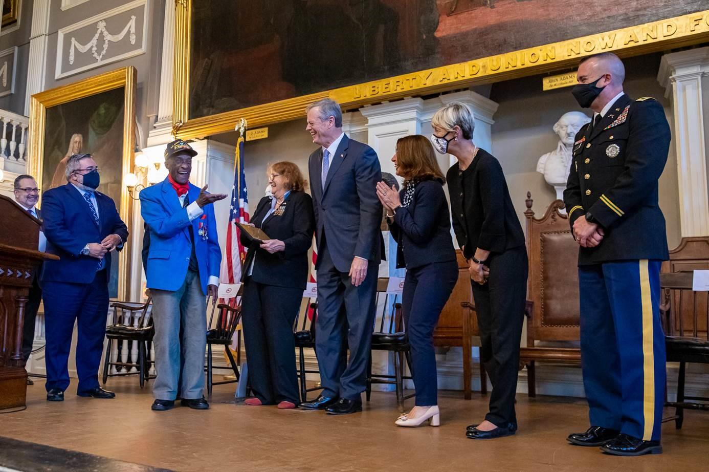 MC Mike Nikitas, Boston Veterans' Services Commissioner Robert Santiago, Lt. Col. Woody Woodhouse, DVS Secretary Cheryl Lussier Poppe, Governor Charlie Baker, Lt. Governor Polito, HHS Secretary Marylou Sudders, and MANG Col. Oberton smile as Lt. Col Woodhouse receives award. 