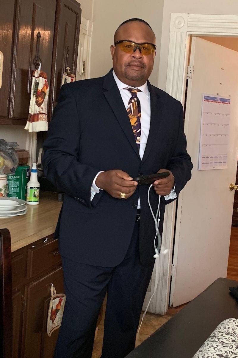 Alvin Calderon stands in a suit, tie, and eyeglasses in an indoor space holding headphones