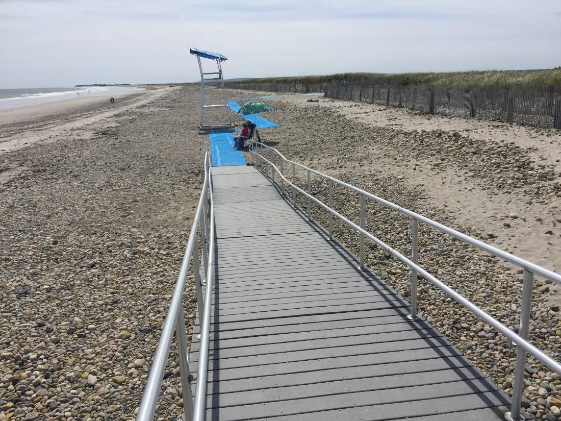 Duxbury - Mobi Mats: Image showing a ramp that runs down to a rocky beach. Where the wooden ramp ends, there are blue Mobi Mats that continue the accessible route to the lifeguard station and another exit/entry point to the beach.