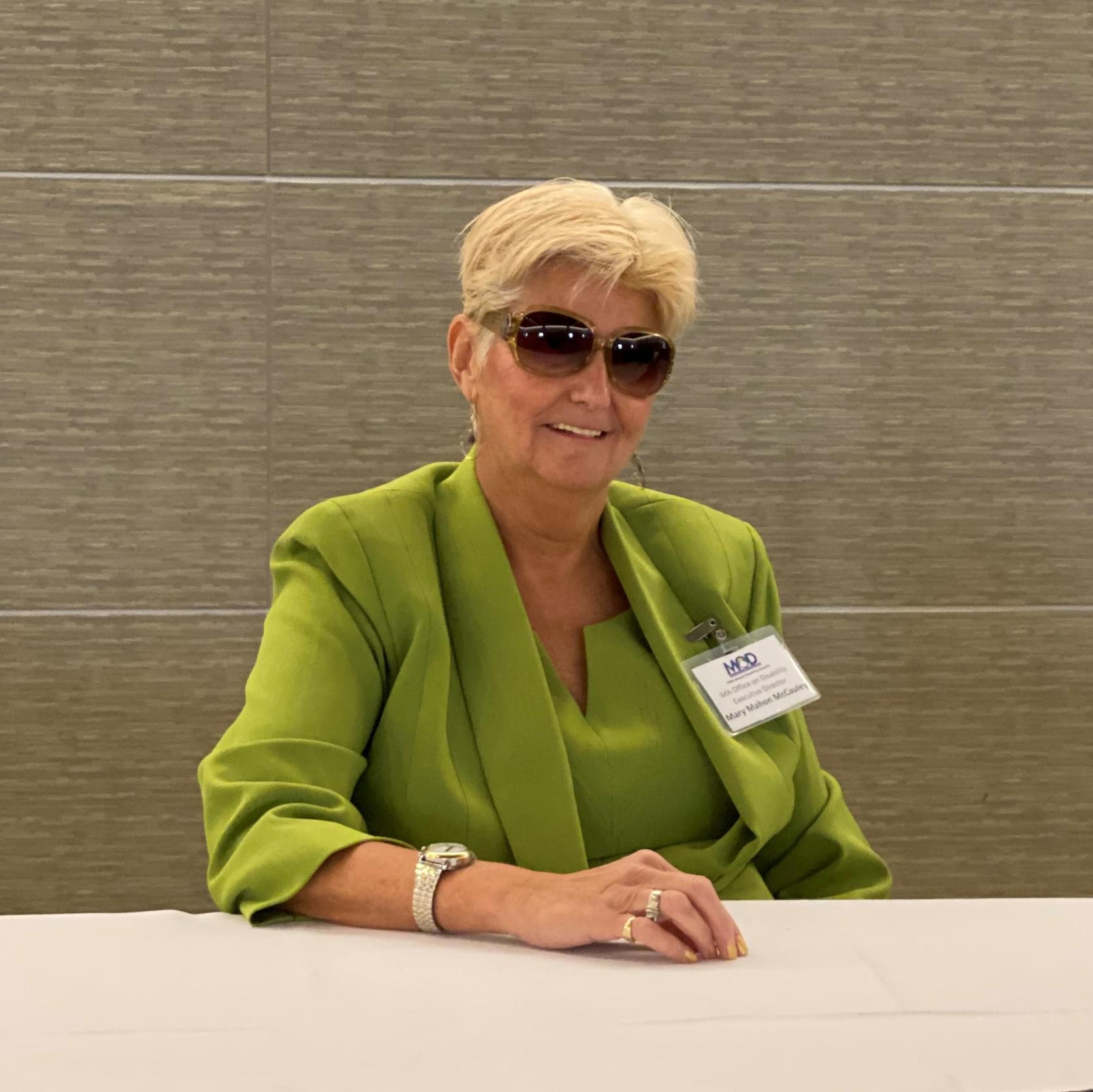 Mary Mahon McCauley, a White woman with short blonde hair, sits at the registration desk at an MOD event. She is wearing a green blouse and jacket. Her right hand is resting on the table and she is wearing a gold watch and rings. She is wearing brown sunglasses and smiling.