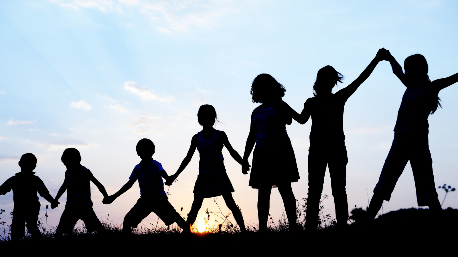 Photo shows sihlouette of children standing in a line and holding hands.