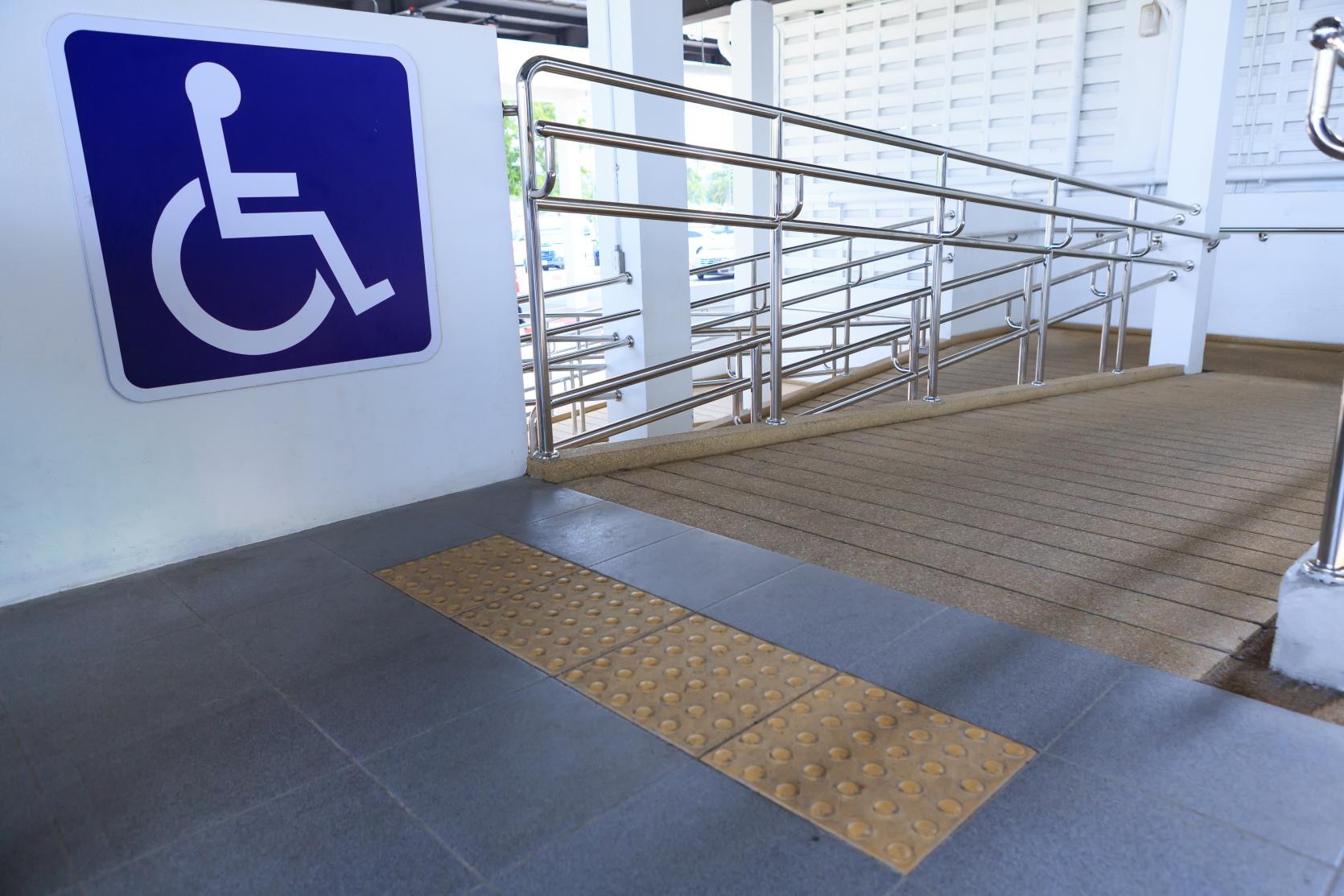 Concrete wraparound wheelchair ramp with stainless steel handrail that comes up to a platform. The platform has a yellow truncated dome surface and a large accessible sign.