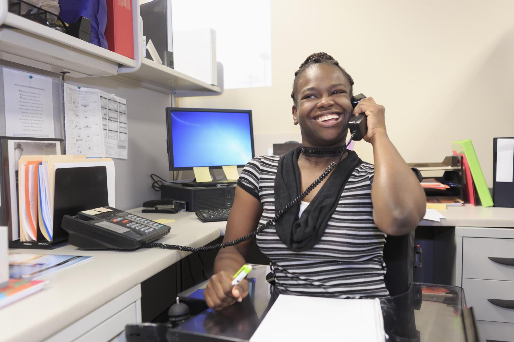 A Black woman using a power wheelchair is sitting at a corner office desk. She is wearing a black and white striped shirt with a black scarf. She is holding a highlighter in her right hand and a desk phone in her right hand, held up to her ear. She has a big smile on her face. The desk is filled with paper files and a desktop computer.