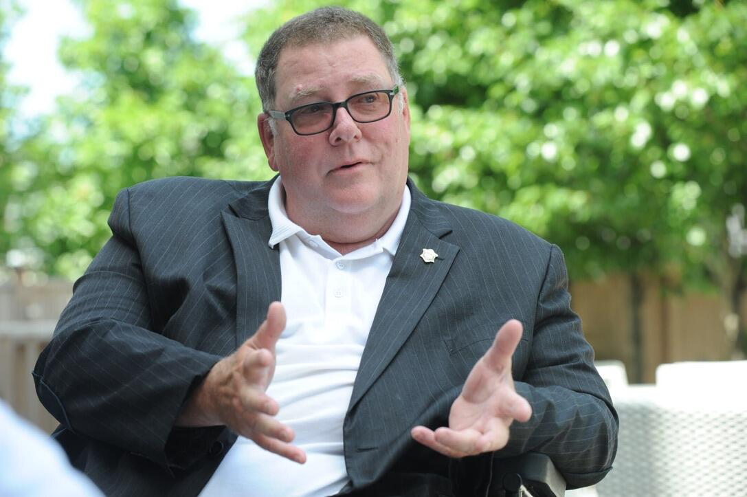 Professional photo of Tom sitting outside, with greenery behind him. He is wearing a gray suit and has his hands out as if he is explaining something