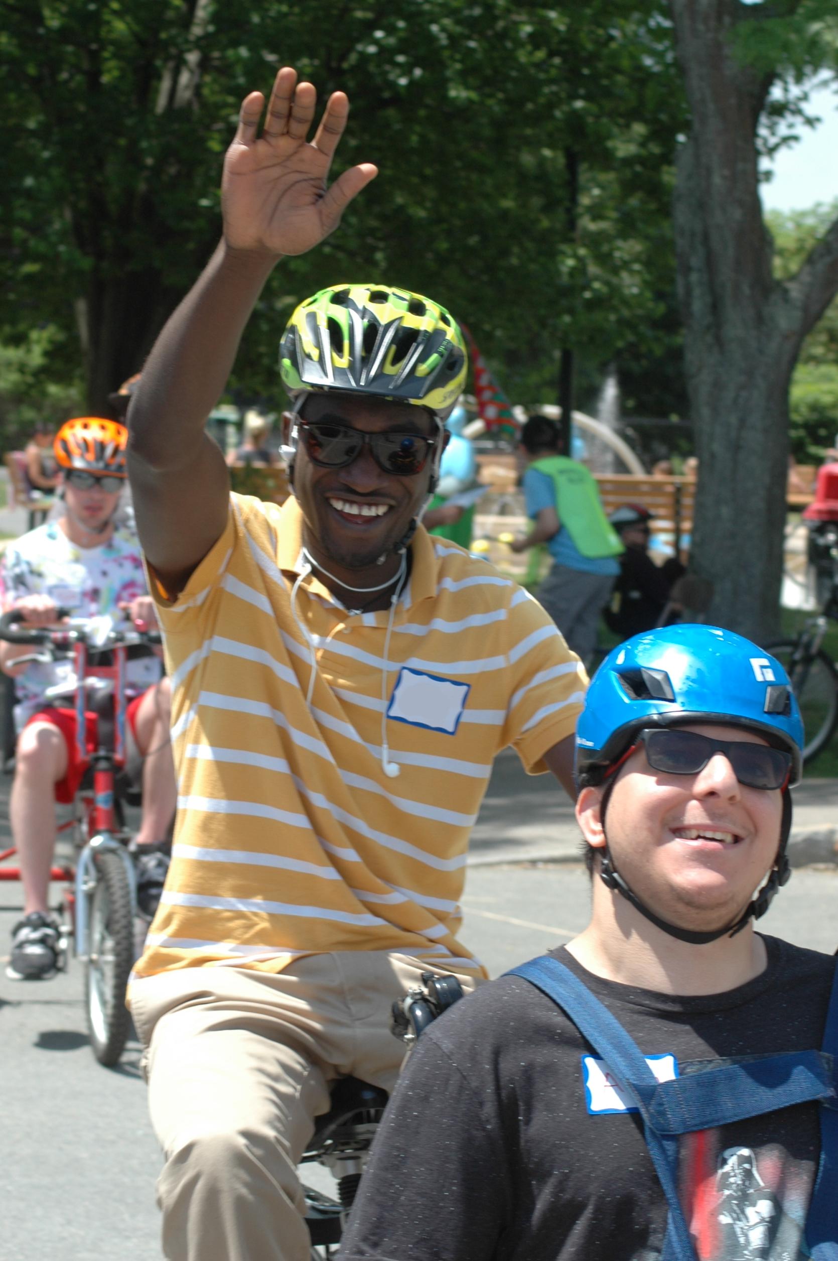 Two smiling cyclists ride by on a tandem cycle and wave at the camera. More cyclists are visible behind them.