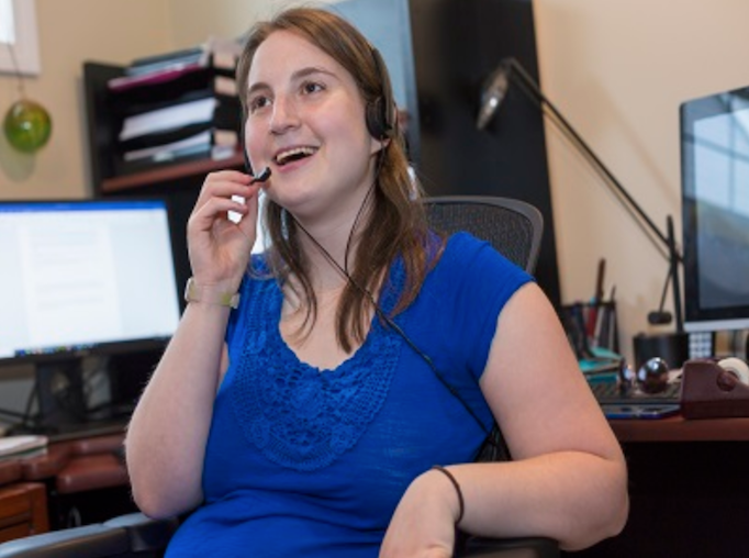 A young white woman with cerebral palsy sits in an office with a headset on. She is turned away from her computer and has a happy look on her face.