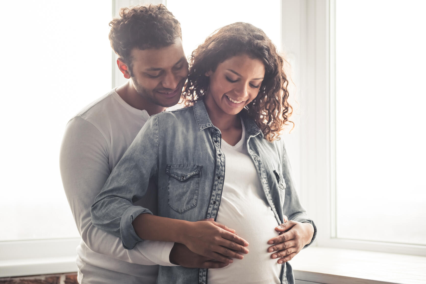 Image of a couple sitting on the couch - one woman who is pregnant, smiling, and the other woman looking lovingly at her partner.