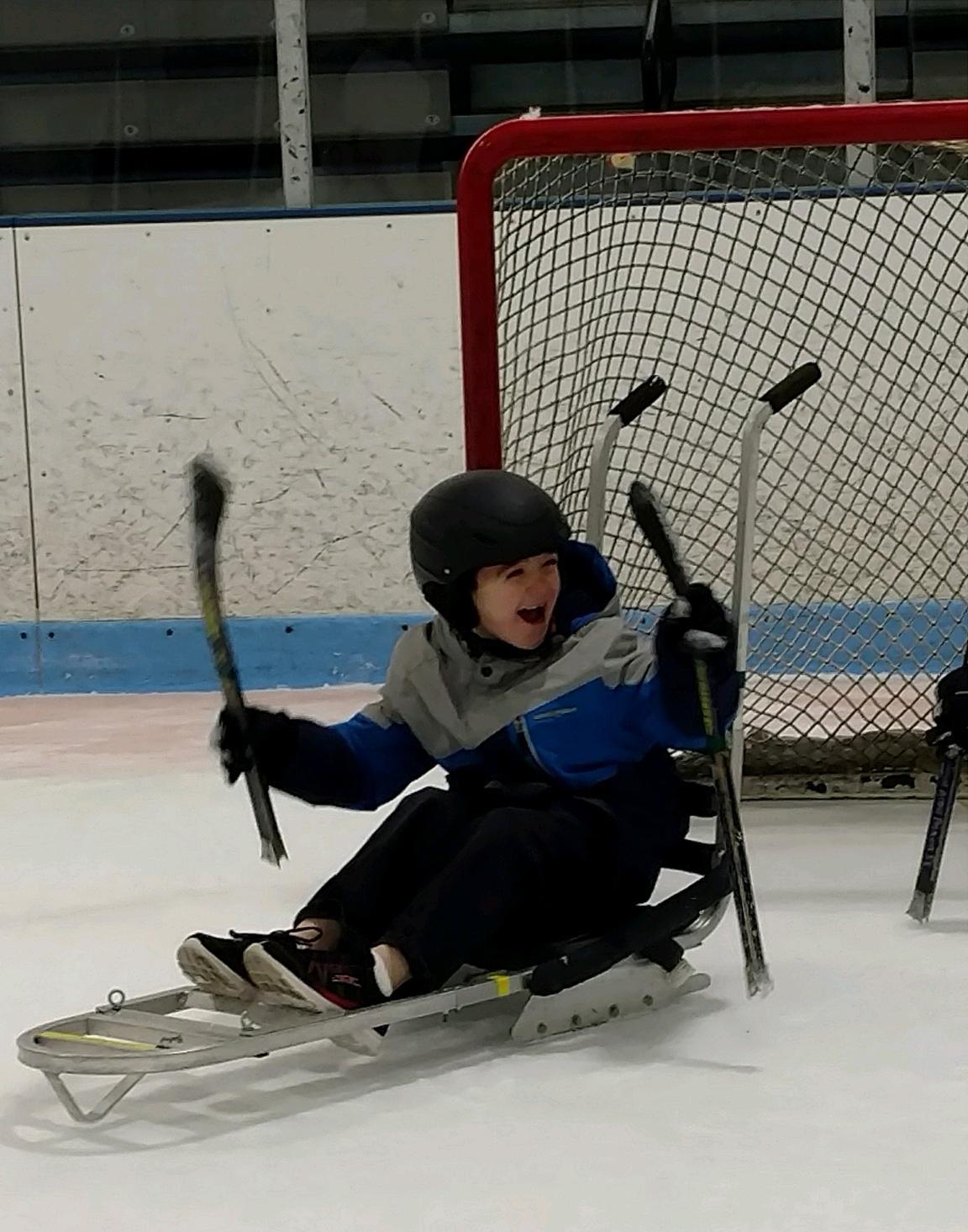 A laughing child in an ice sled in front of a hockey net.