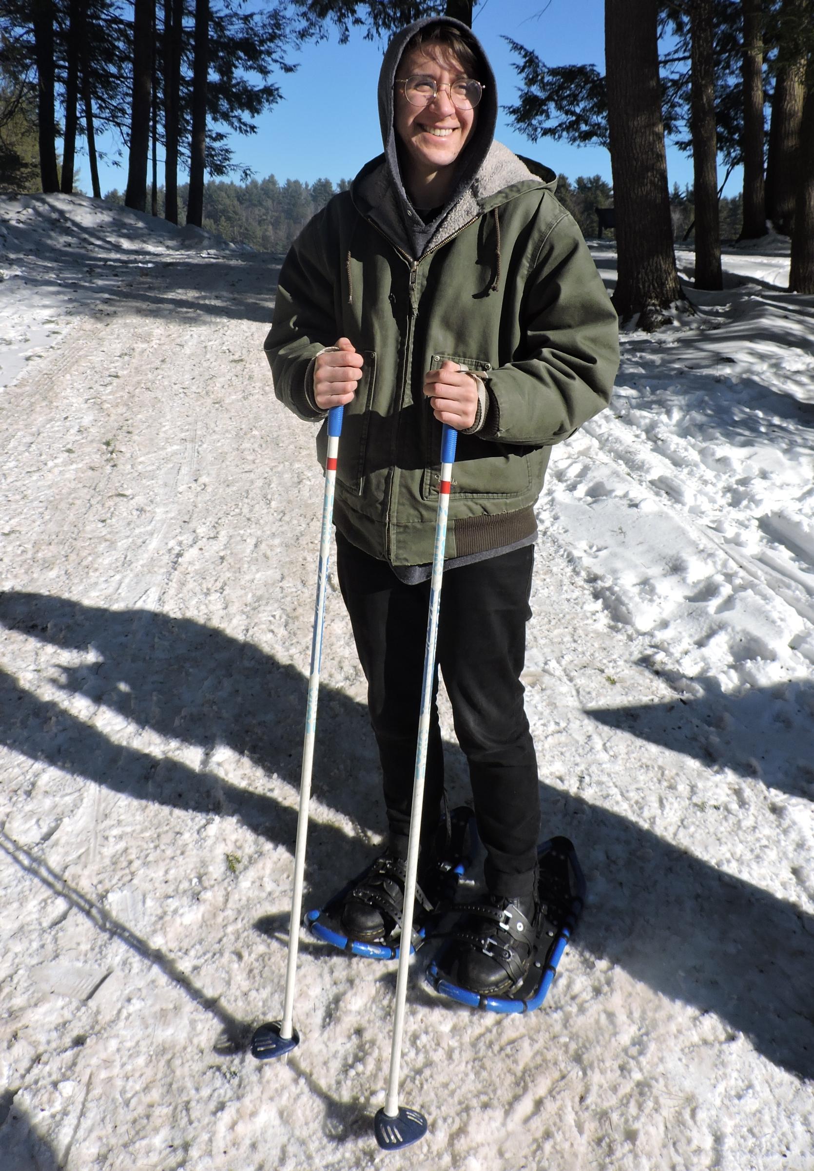 A person wearing snowshoes and holding ski poles is standing at the foot of a snowy trail smiling at the camera.