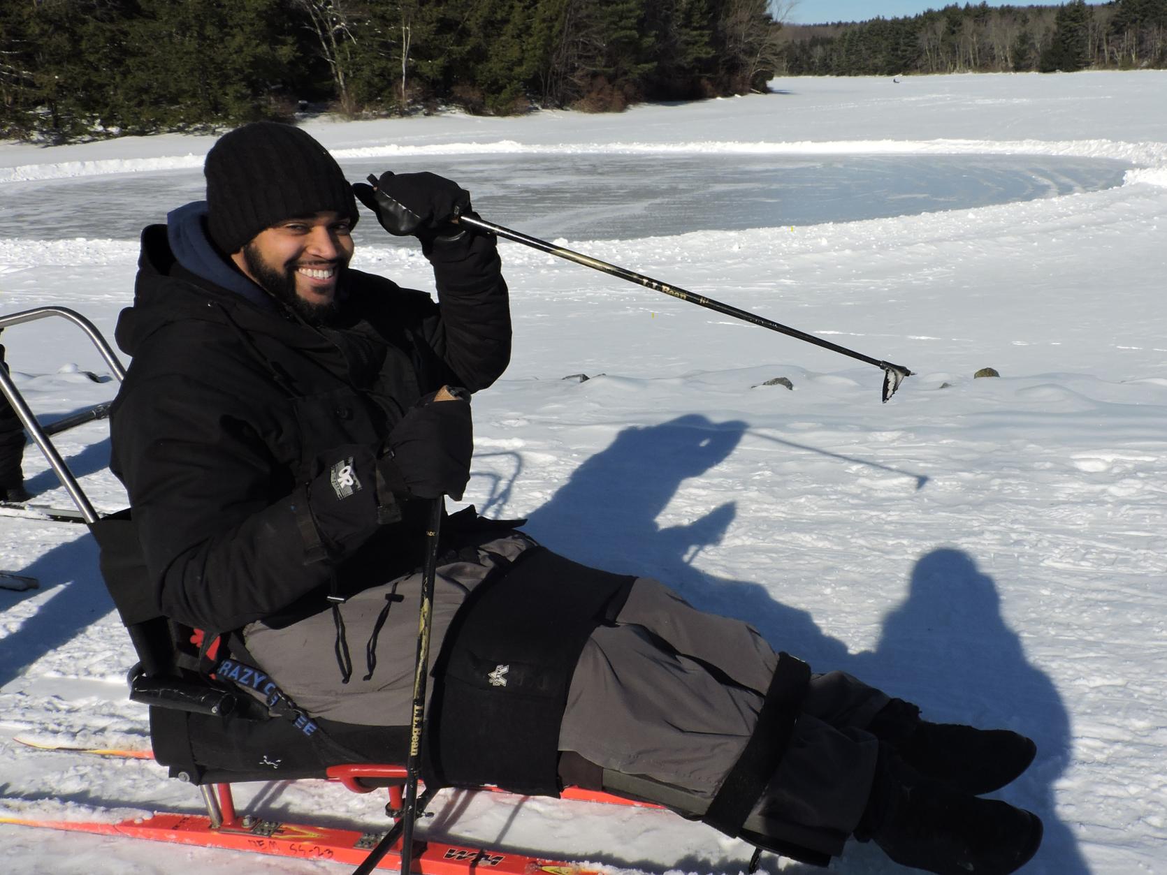A smiling sit-skier in the snow next to a frozen lake.