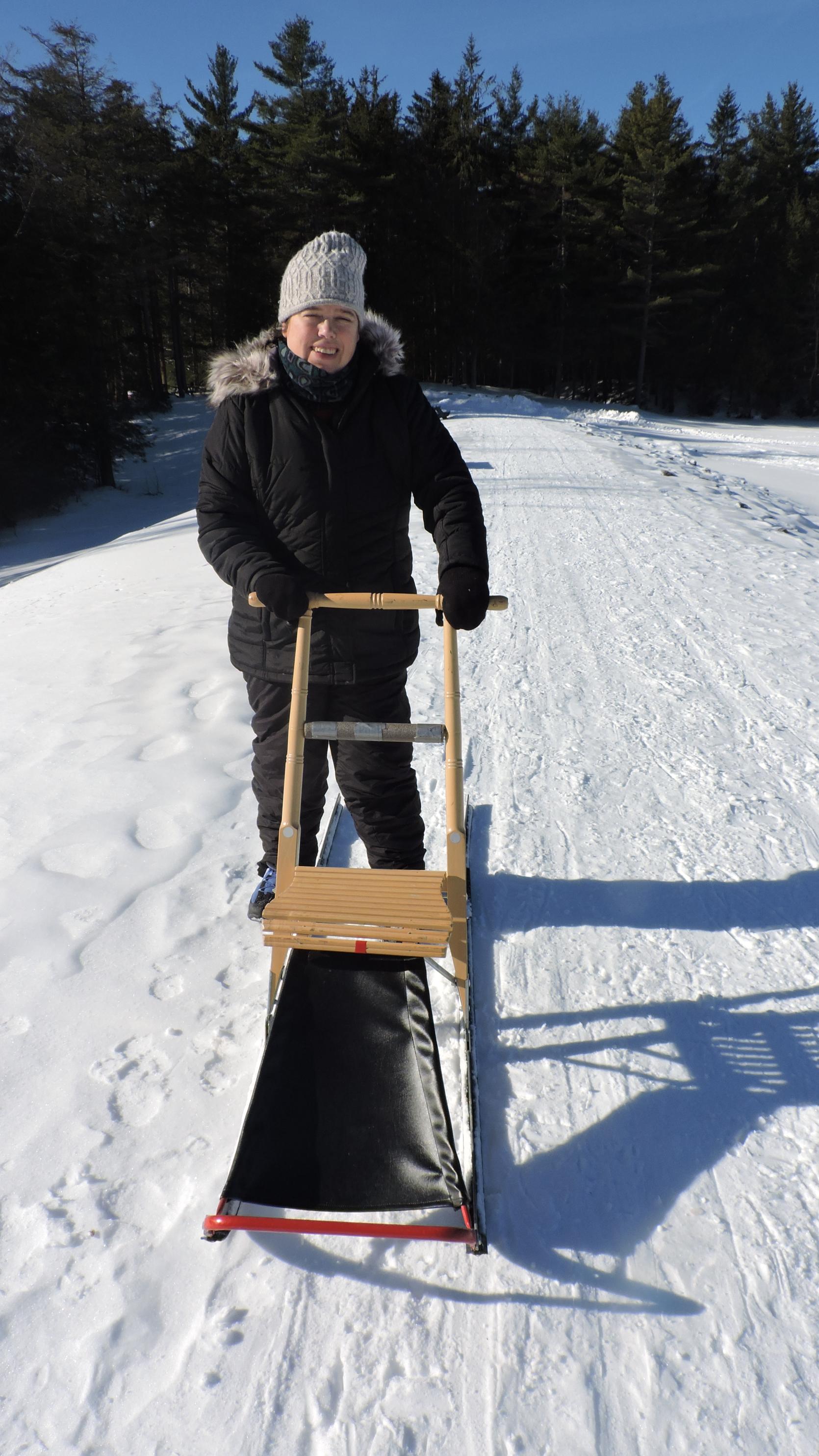 A person standing behind a kick sled on a snowy trail on a sunny day.
