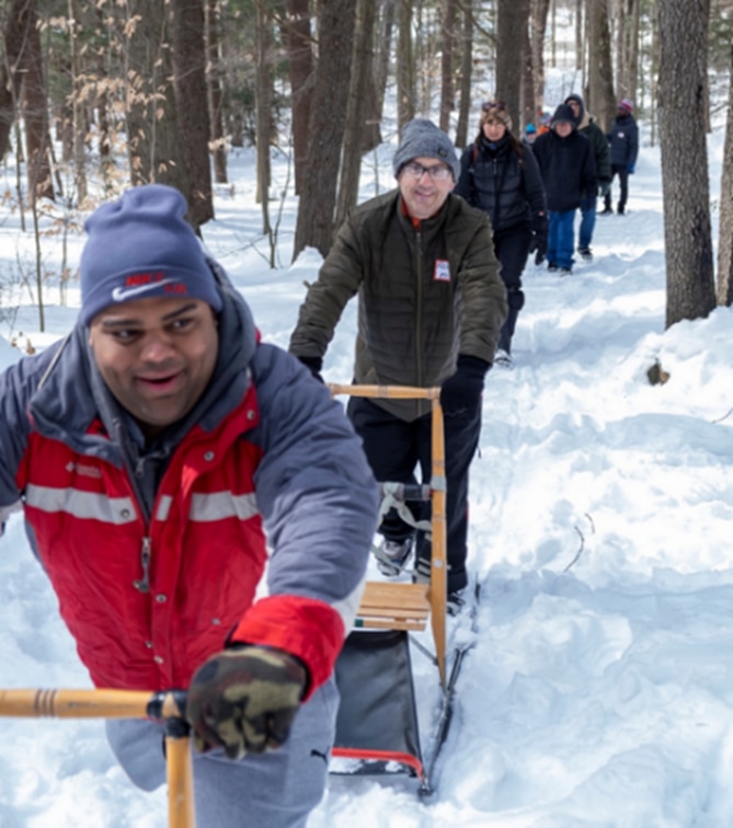 A group of hikers on a snowy trail in the woods. Two hikers are pushing kick sleds.