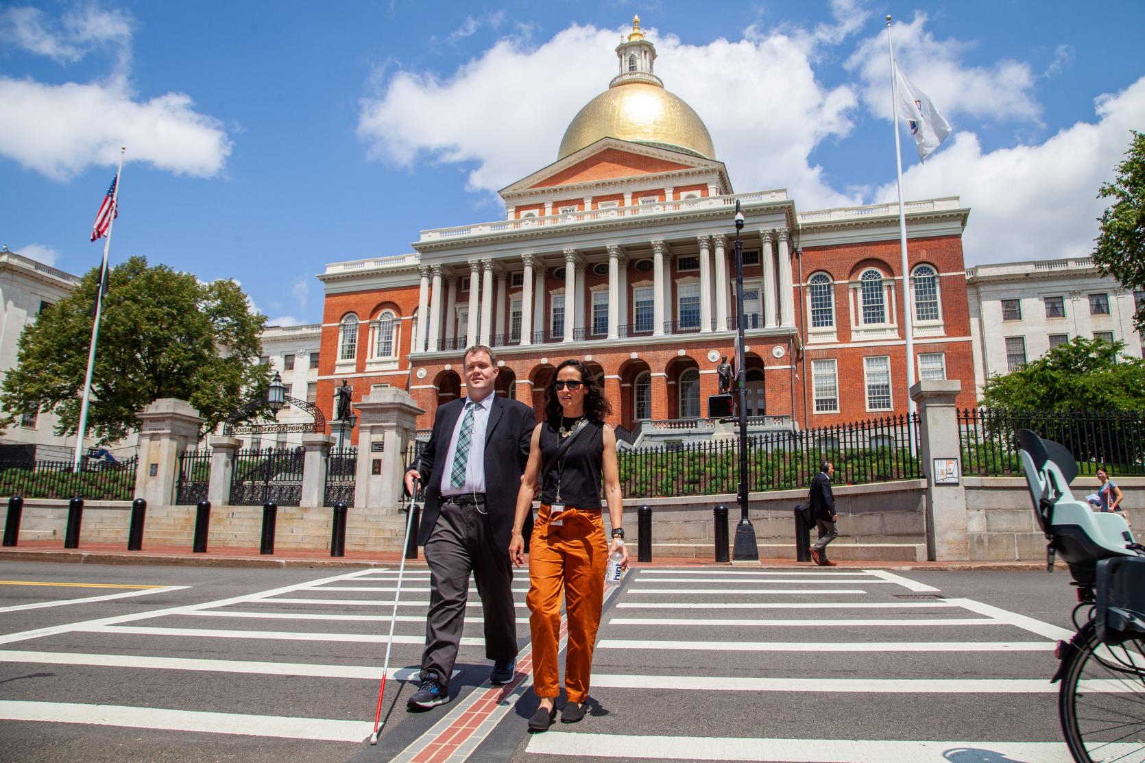 A photo of a man using his white cane crossing in a crosswalk in front of the Massachusetts State House