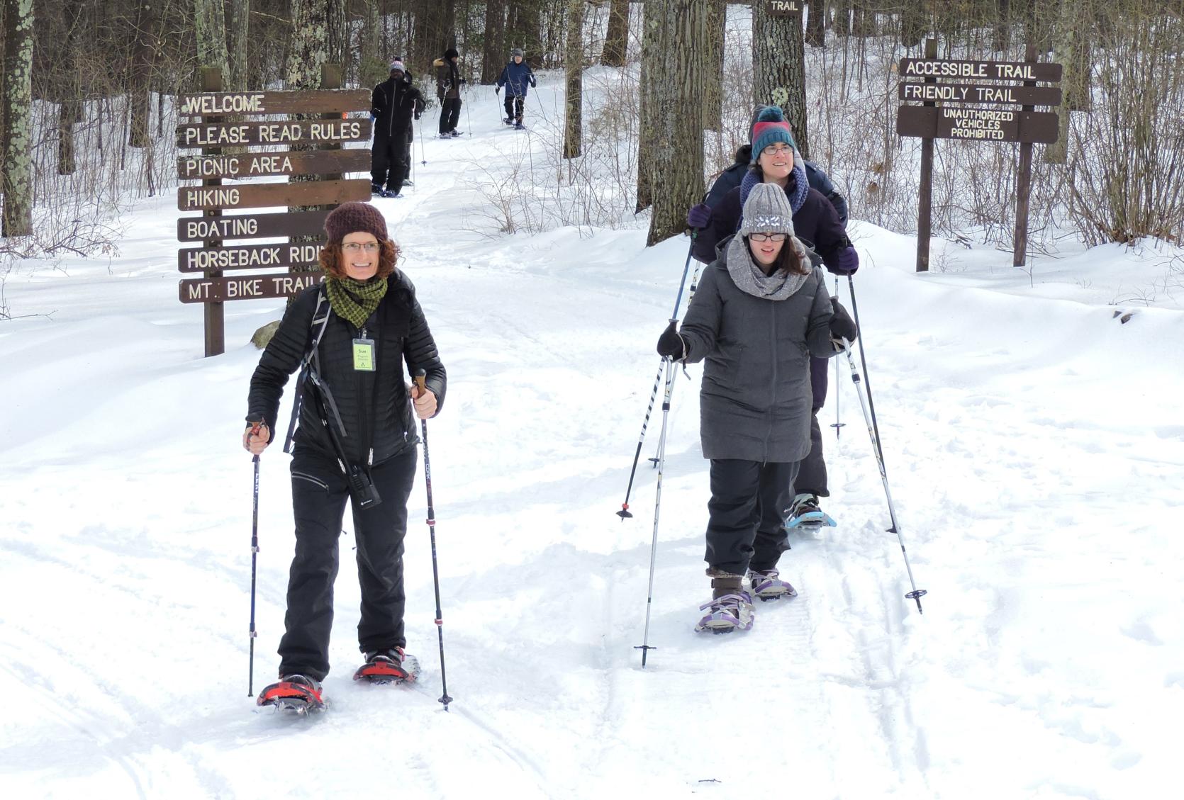 Snowshoers using ski poles on a snowy winter trail in the woods.