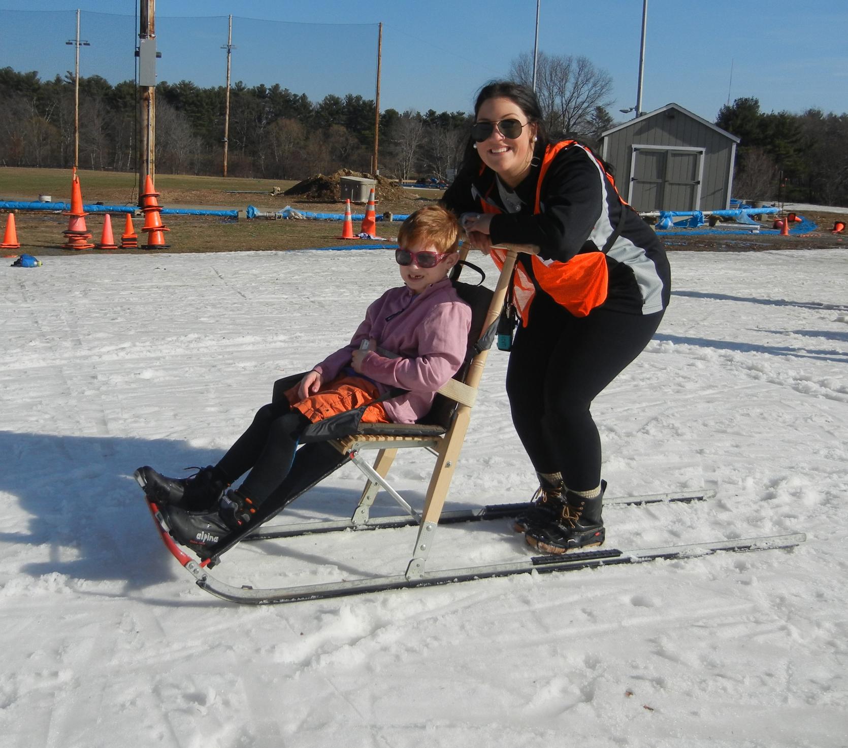 A young skier on a kick sled on a groomed trail with a volunteer standing behind them.