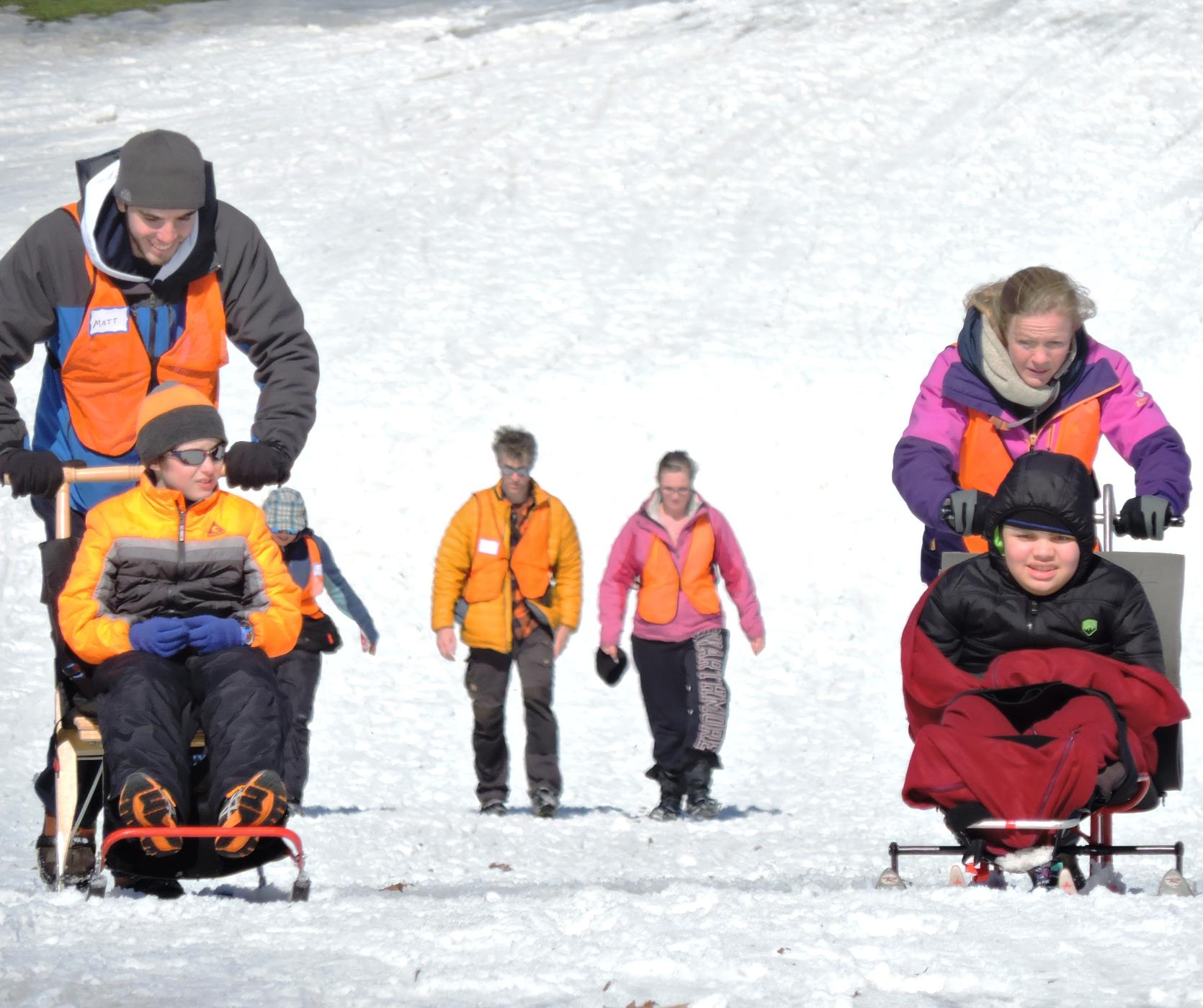A kick sledder and a sit-skier are being pushed down the hill by volunteers. Behind them, two other people are walking down the hill wearing ice grippers on their shoes.