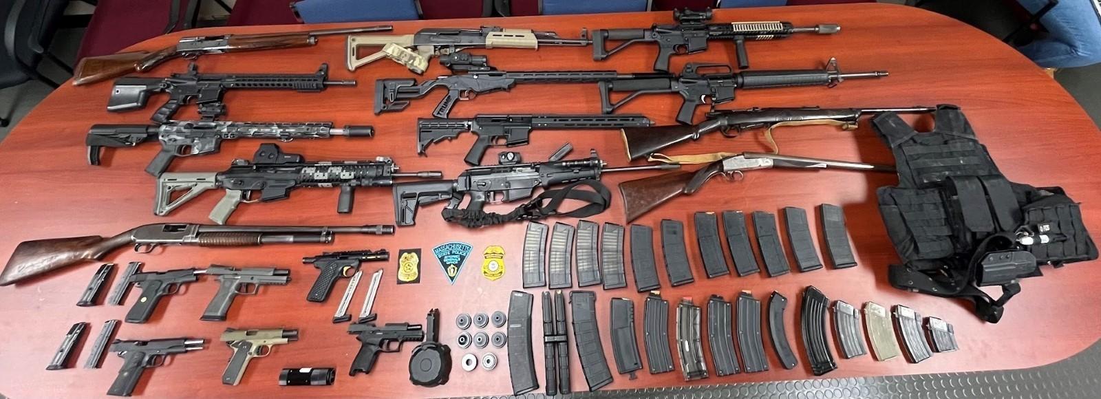 Guns on the table include: 7 AR-15 style rifles, 1 AK-47 rifle, 1 .22 bolt action rifle, 3 shotguns, 1 .303 bolt action rifle, 8 handguns, 23 large capacity magazines capable of holding more than 10 rounds of ammunition, 2 silencers, and hundreds of rounds of various caliber ammunition