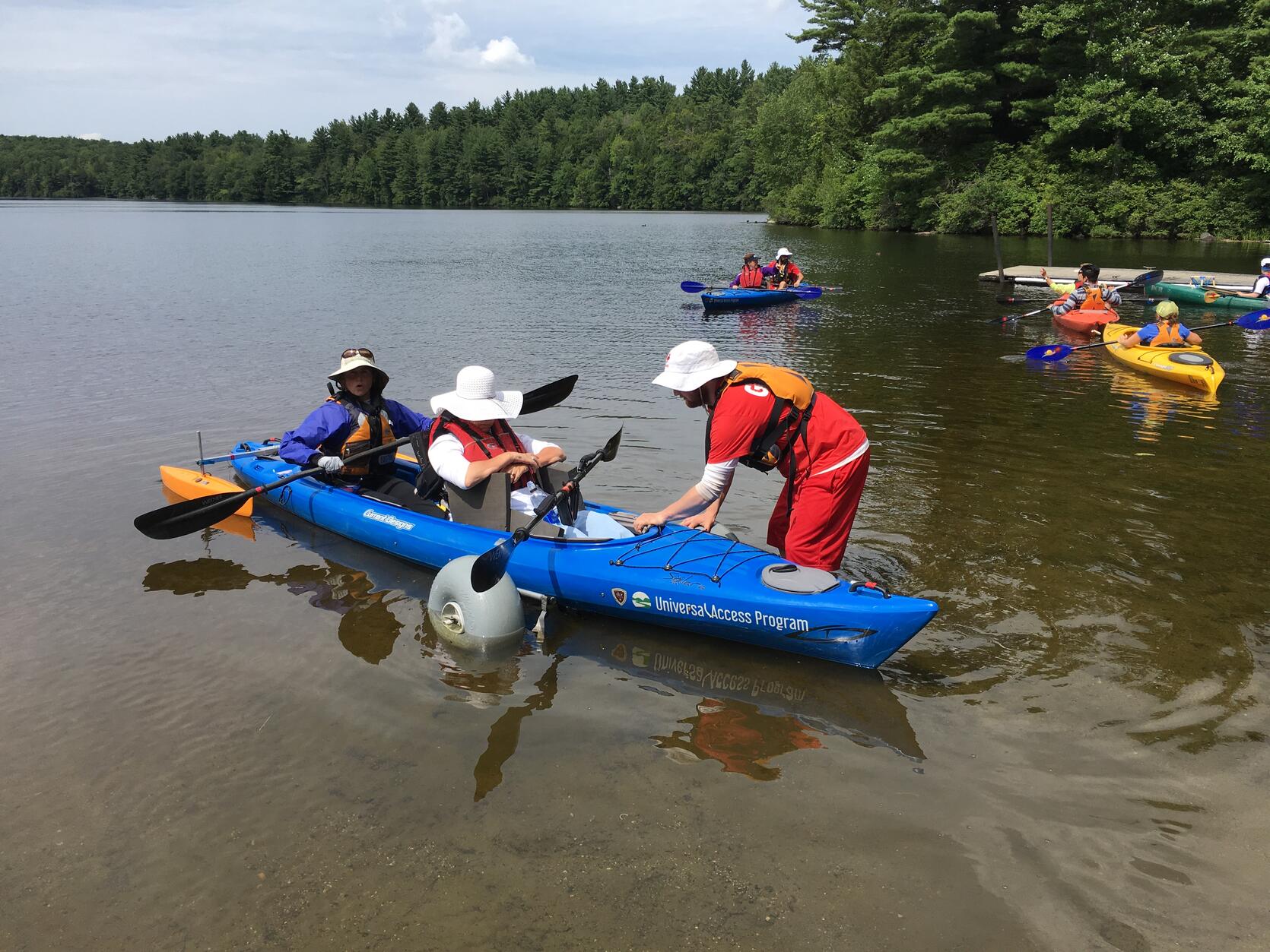 Two people sitting in a tandem kayak equipped with outriggers, are holding paddles as they are wheeled into the reservoir. 