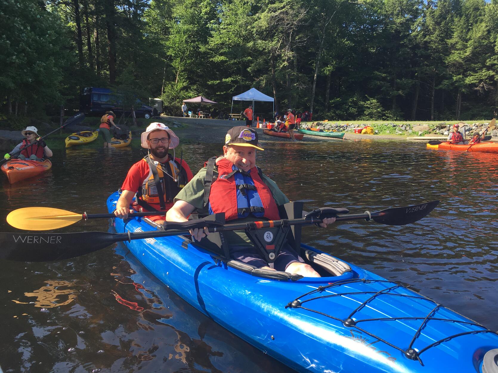 Two people are smiling in a tandem kayak as they head out from shore on a tree-line lake.  