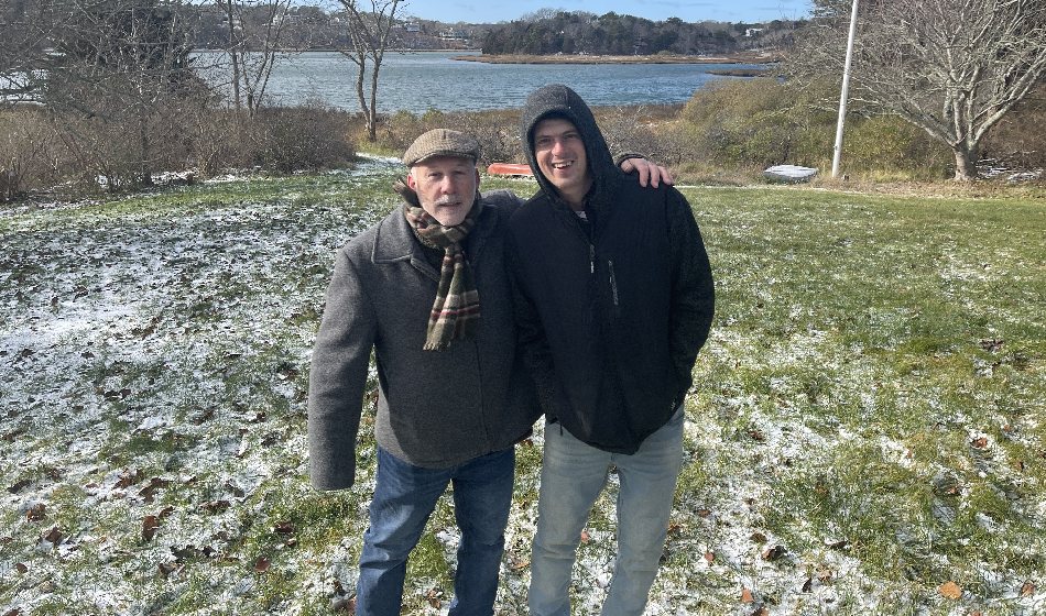 Two men stand on a hill overlooking a body of water, posing for a photo