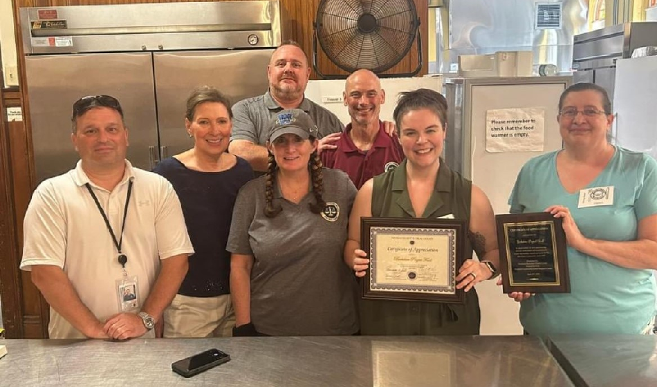 A group poses for a picture in a kitchen. The two right-most women hold framed certificates.