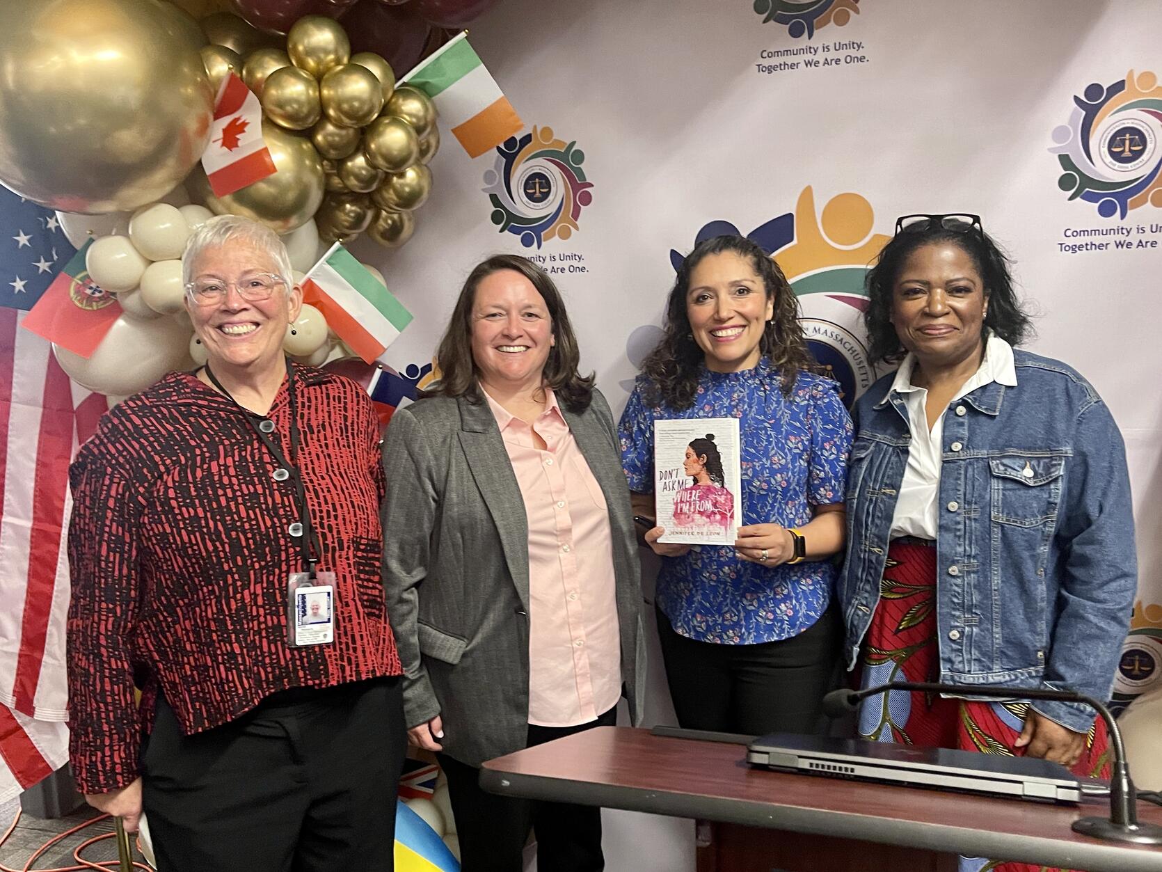Four women standing together with balloons and international flags in the background. One woman is holding a book titled "Don’t Ask Me Where I’m From."