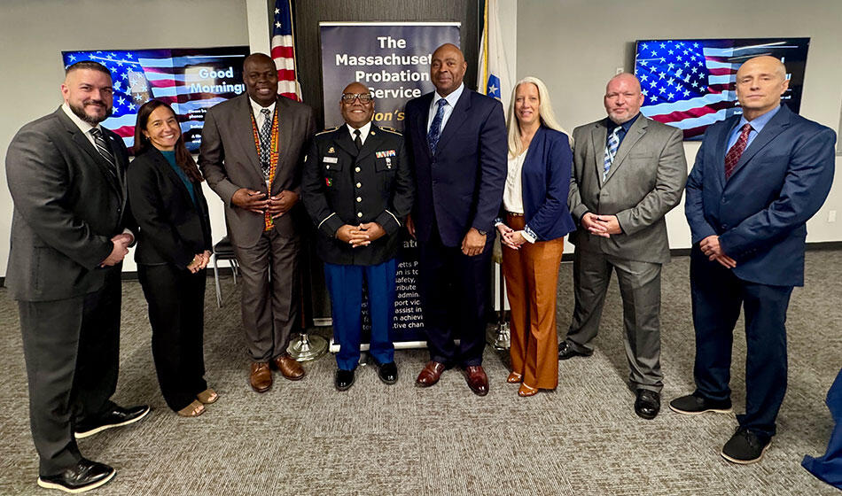 A group of eight people stand in front of the Massachusetts Probation Service banner and two American flags indoors.