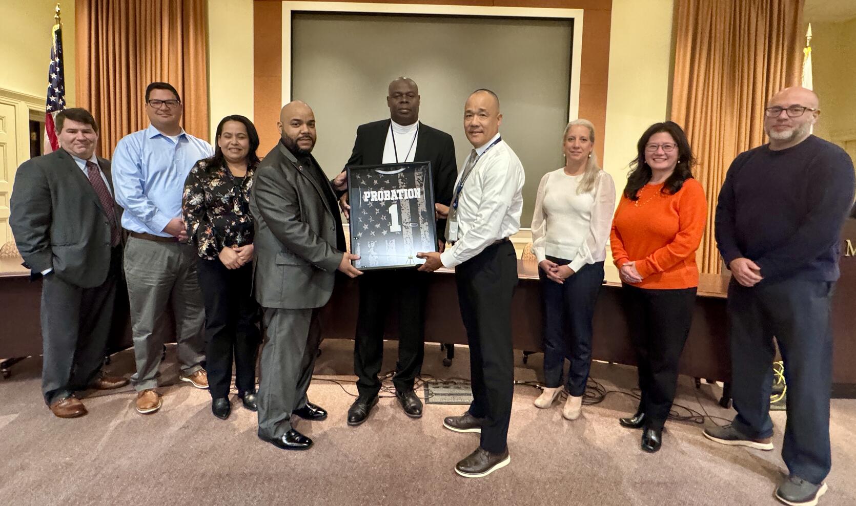 A group of people pose for a picture with the middle-most people holding an encased probation basketball jersey.