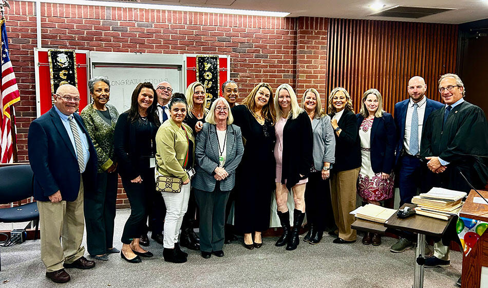 A group of people, including a judge, stands in a courtroom setting with "Congratulations" decor on a brick wall.