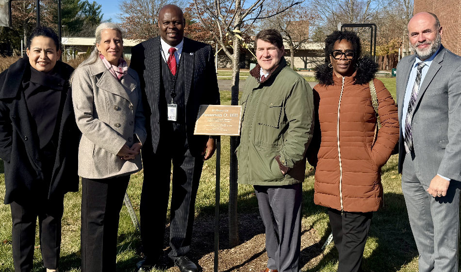 Six people pose with a plaque in-between them flanked by a newly planted tree.