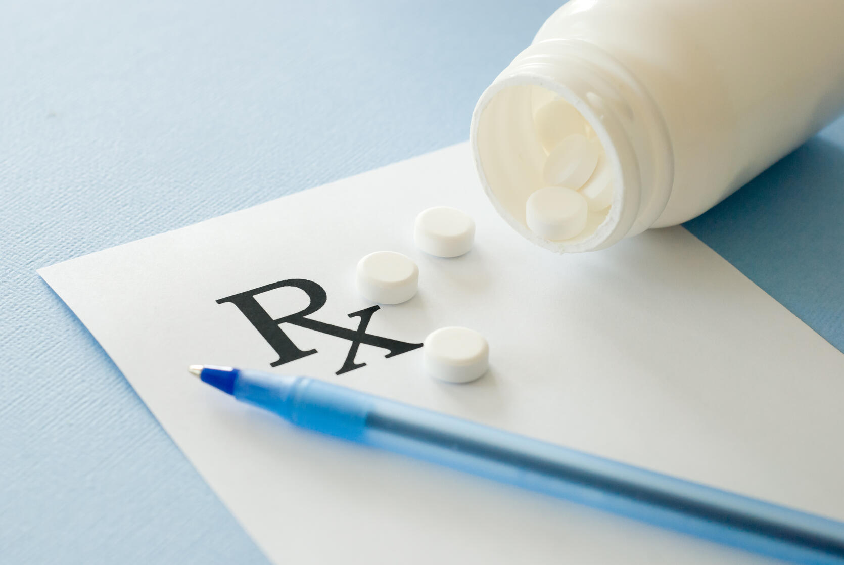 Prescription pad with pen on a table. Pills spill out from a bottle onto the pad.