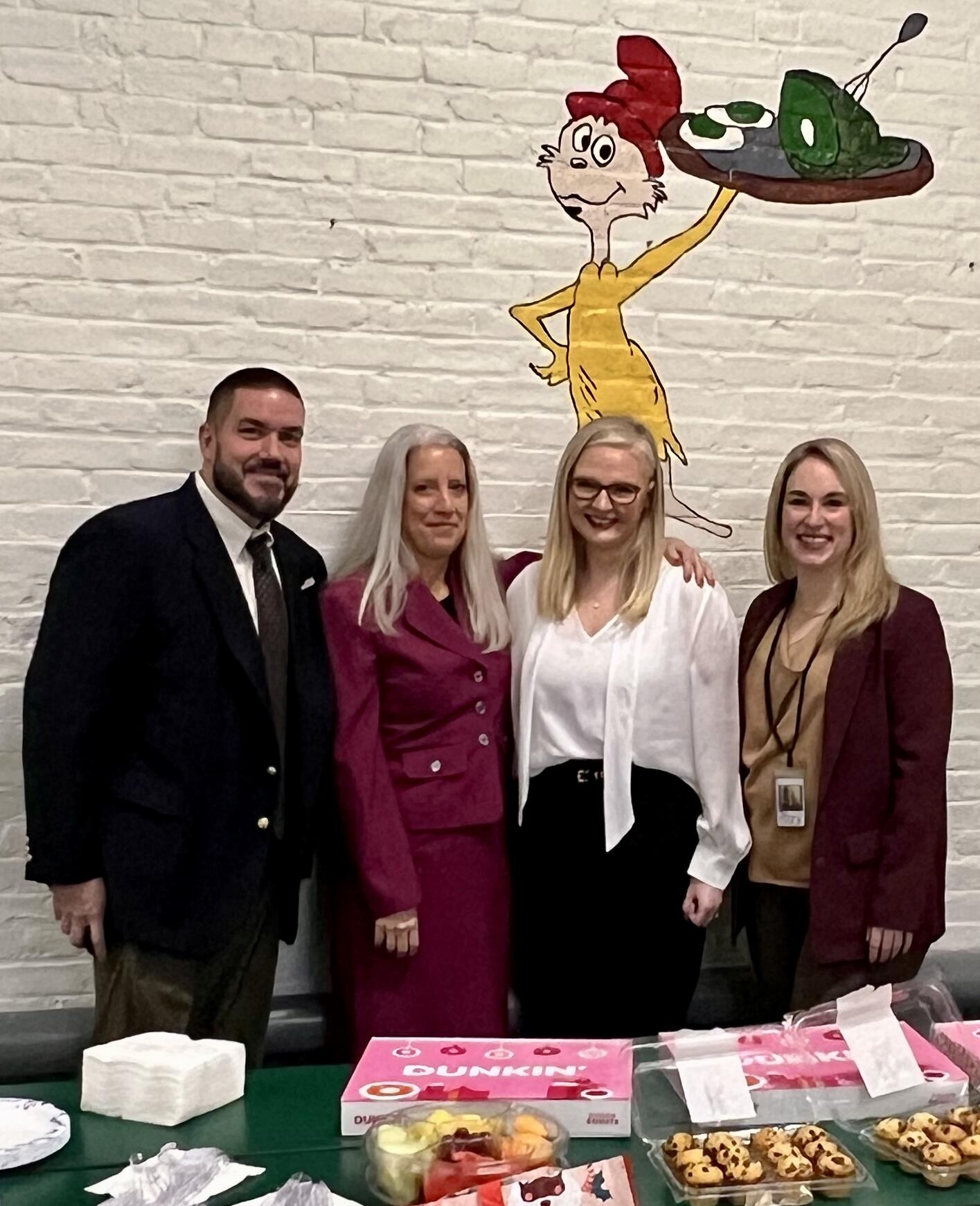 Four people standing in front of a cartoon mural of a character with a table of food in front of them.