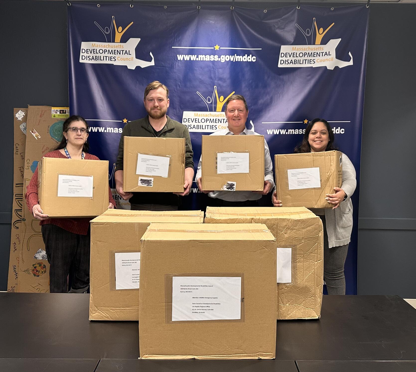 Four staff members standing in front of an MDDC banner with seven shipping boxes.
