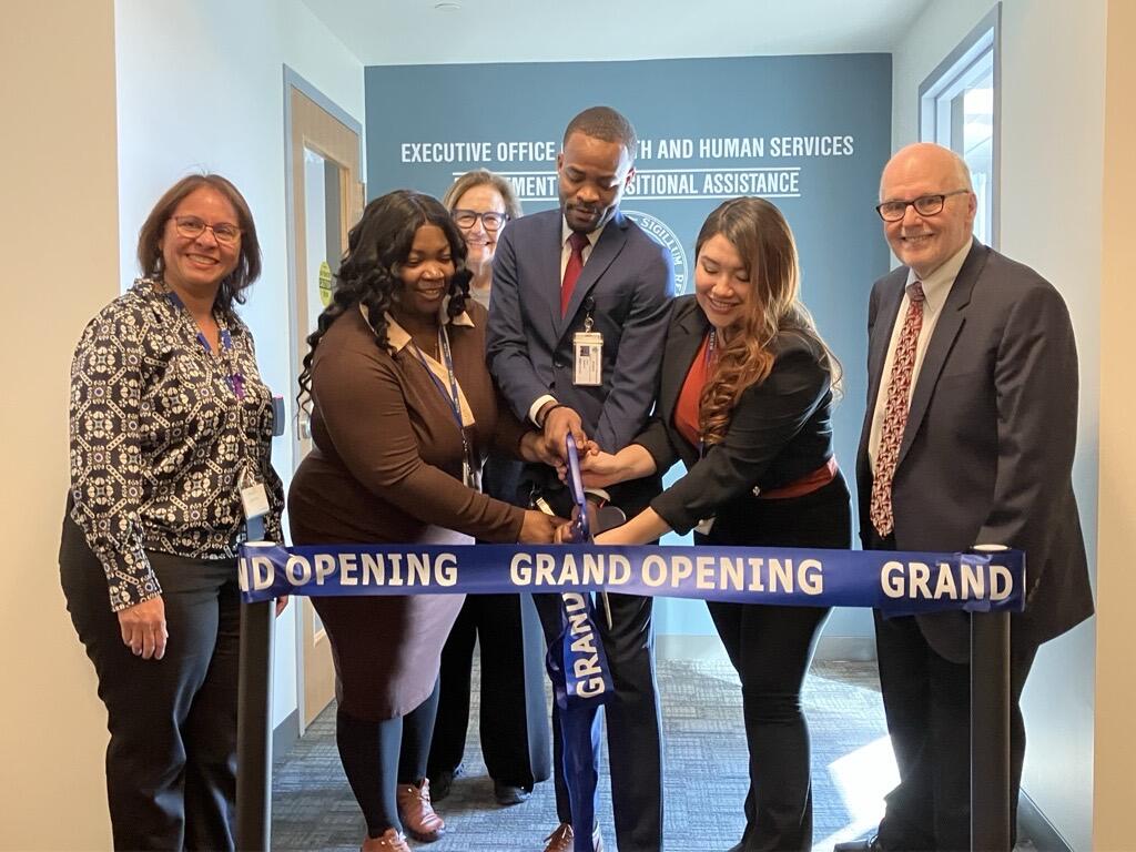 DTA Jackson Square office Director Patria Espinal (left), Front-End Staff Cindy Saintus, Jeff Saintus, and Ana Castrillon (center), HHS Secretary Kate Walsh (back), and  DTA Commissioner Jeff McCue (right) cut ribbon outside the lobby doors of the new DTA Jackson Square Office