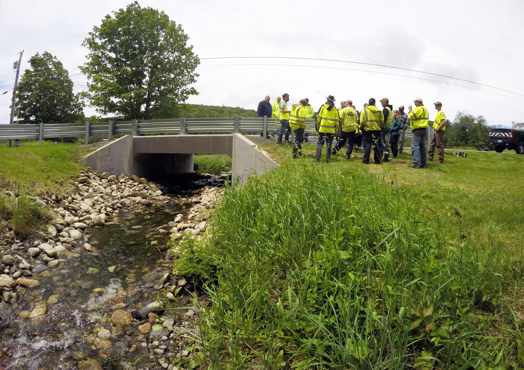 A group of municipal staff stand behind a road barrier next to a large culvert.
