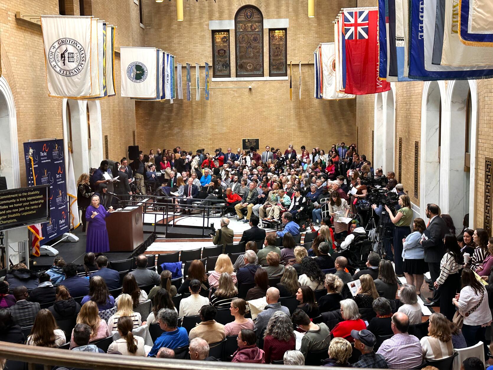 An audience of people inside of the State House's Great Hall of Flags.