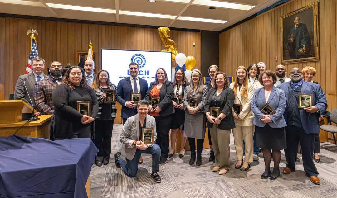 A group of 18 people holding plaques in a conference room, posing in front of a screen displaying "E.A.C.H Emerging Adult Court of Hope."